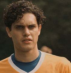 a young man with curly hair wearing an orange and blue soccer jersey looking at the camera