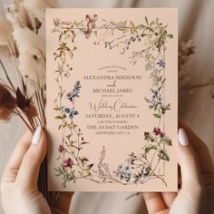 two hands holding up a wedding card with flowers on it and greenery in the background