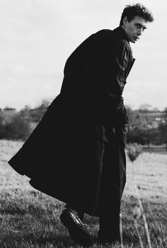 black and white photograph of man in trench coat leaning on pole with grass field behind him