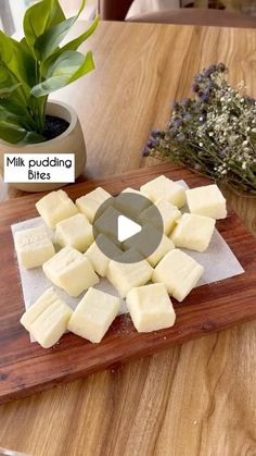 a wooden cutting board topped with cheese on top of a table next to a potted plant