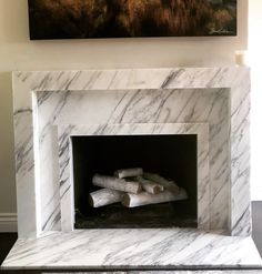 a white marble fireplace with some logs in the middle and a painting on the wall above it