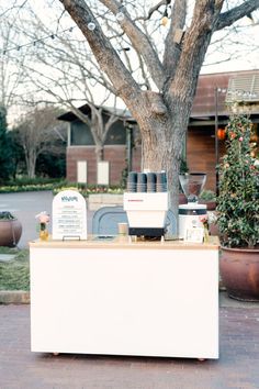 a white counter sitting next to a tree on top of a brick floored sidewalk