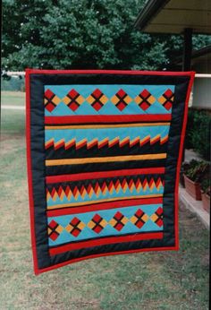 a blue and red quilt hanging from a clothes line in front of a house with trees