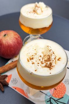 two glasses filled with drinks sitting on top of a table next to an apple and cinnamon stick