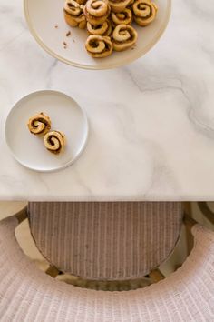 some food is sitting on a white plate near a bowl of cinnamon buns and a pink chair