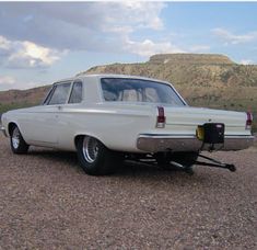 an old white car parked in the middle of a dirt field with mountains in the background