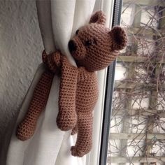 a crocheted teddy bear hanging from the side of a window sill next to a white curtain