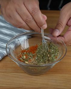 two hands are mixing spices in a glass bowl on a wooden counter top, with another person's hand holding a metal spoon