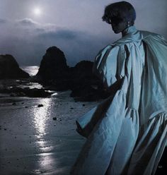 a woman standing on top of a sandy beach next to the ocean under a cloudy sky