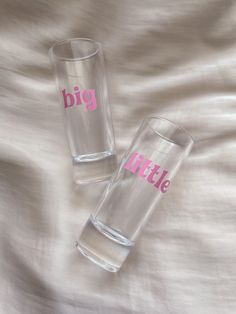 two clear glasses with pink lettering sitting on top of a white bed sheet next to each other