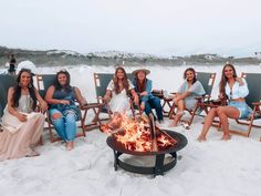 a group of women sitting around a fire pit on the beach with drinks in their hands