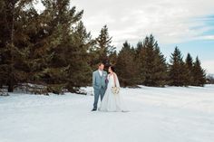 a bride and groom standing in the snow near some pine trees at their wedding day