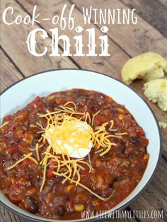 a white bowl filled with chili and cheese on top of a wooden table next to bread