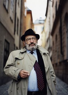 a man in a trench coat, tie and hat standing on an alleyway with buildings behind him