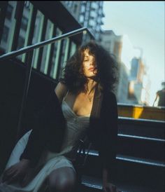 a woman sitting on some stairs in the city at sunset or dawn, wearing a white dress and black blazer