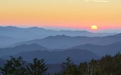 the sun is setting in the mountains with trees on the foreground and hills to the far side