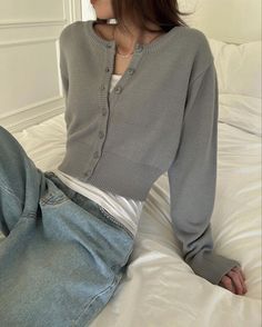 a woman sitting on top of a bed wearing a gray cardigan sweater and jeans