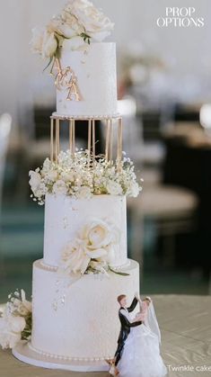 a wedding cake with white flowers and gold letters on the top tier is displayed in front of a table