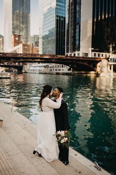 a bride and groom standing next to each other near the water in front of tall buildings