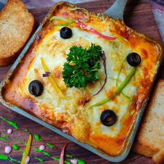 a square pizza with olives, cheese and bread on a cutting board next to it