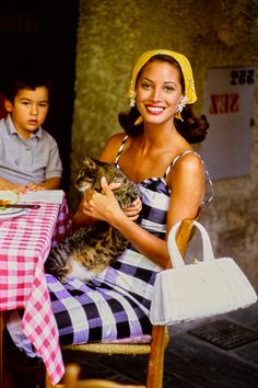 a woman holding a cat while sitting at a table with a boy in the background