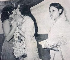 two women standing next to each other in front of a white wall and one woman is kissing the man's cheek