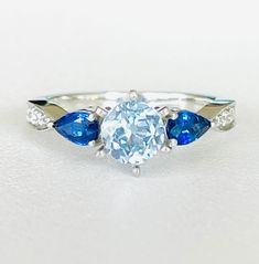 Stunning engagement or birthstone ring! This gorgeous ring features a light blue, 5.5mm, 0.75ct, round, natural topaz. The topaz is flanked by two, pear cut, royal blue, 5x3mm, 0.6ctw sapphires. The band has 10, colorless, eye-clean, 0.11ctw, natural diamonds. The six prong setting and pinched band are crafted in platinum. The ring weighs 5.3 grams. The band is 2.75mm at its widest point. The ring is size 5.5 and sizeable by a jeweler. This gorgeous ring is in excellent condition! Unique Engagement, Pear Cut, Birthstone Ring, Ring Verlobung, Prong Setting, Natural Diamonds, Topaz, Royal Blue, Pear