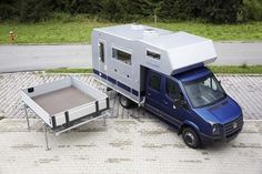 a small blue van parked next to a table on the side of a road with an awning over it