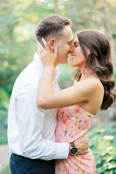 a man and woman standing close to each other in front of some trees with their arms around each other