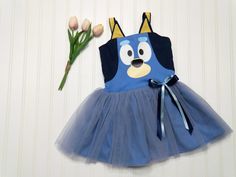 This kids Blue Heeler character dog dress is a charming and playful outfit perfect for little ones. The dress features a vibrant shade of blue, adding a pop of color to any occasion.  It has a character face and ears adorning the bodice.  The skirt has tulle over the cotton fabric adding a touch of whimsy and fun.  The back of the bodice has elastic encased for ease of dressing and will stretch for extended wear. Also available is little sister Orange Heeler. Please select Orange or Blue in the dropdown for your desired character. Sizes available are from 6 months to size 6. *Please allow two weeks from your date of order for your dress to ship.* Blue Disney Dress For Costume Party, Blue Fitted Disney Dress, Whimsical Blue Dress For Costume Party, Whimsical Blue Dresses For Costume Party, Whimsical Blue Costume Dresses, Cute Blue Cartoon Print Dress, Cute Blue Costume Dress, Cute Blue Dress With Cartoon Print, Playful Blue Dress For Playtime