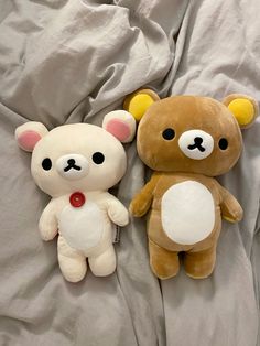 two teddy bears laying on a bed with white sheets and grey sheets, one is brown and the other is tan