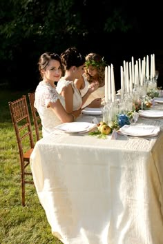 three women sitting at a table with white plates and candles on it in the grass