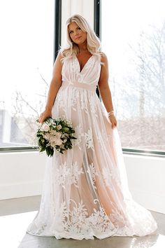 a woman standing in front of a window wearing a wedding dress and holding a bouquet