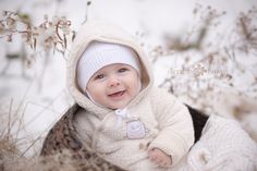 a baby is smiling while sitting in the snow wearing a white coat and hat with a bow on it's head