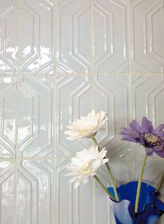 three white and purple flowers in a blue vase against a wall with geometric tiles behind them