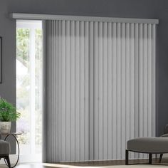 a living room with white vertical blinds in the window, and a grey chair next to it