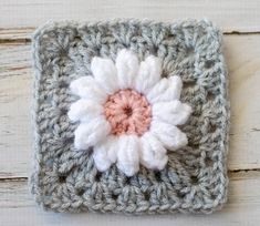 a crocheted square with a white and pink flower on the center is sitting on a wooden surface