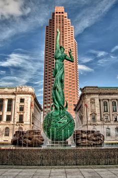 there is a green statue in the middle of this square with buildings behind it and blue skies above