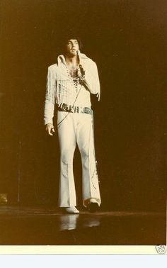 a man standing on top of a stage with a microphone in his hand and wearing white pants