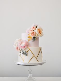 a three tiered cake with pink and white flowers on top, sitting on a glass pedestal