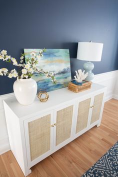 a vase with flowers on top of a white cabinet next to a painting and lamp