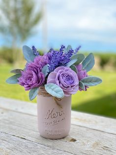 purple flowers are in a mason jar on a wooden table with grass and sky in the background