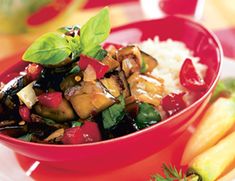 a red bowl filled with vegetables and rice on top of a table next to fruit