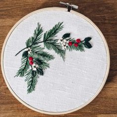 a cross stitch pattern with pine needles and red berries on white linen in a wooden hoop