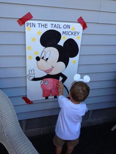 a little boy is painting a mickey mouse sign on the side of a house that says pin the tail on mickey