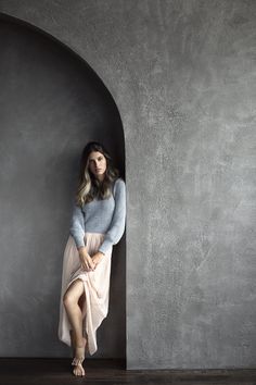 a woman is leaning against a wall with her legs crossed and wearing a long skirt