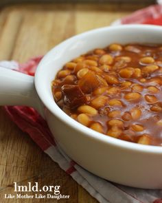 a white bowl filled with baked beans on top of a wooden table next to a red and white checkered napkin