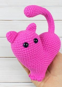 a hand holding a pink crocheted cat ornament in front of a white wooden background