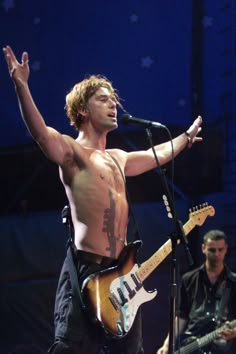 a man with no shirt on singing into a microphone and holding his guitar in the air