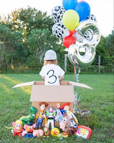 a cardboard box with balloons and toys in it on the grass next to a birthday cake